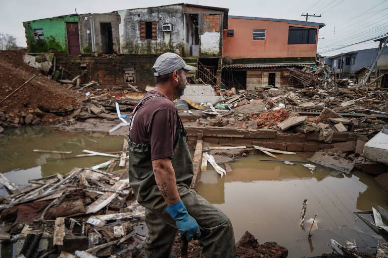 A imagem mostra um homem de boné e luvas azuis observando a destruição causada por um desastre natural. Ele está cercado por escombros, madeira quebrada e água acumulada. Ao fundo, há casas danificadas, uma delas com uma fachada verde e outra com uma fachada laranja.