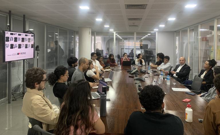 A imagem mostra uma reunião em uma sala de conferência com várias pessoas sentadas ao redor de uma mesa retangular de madeira. Algumas pessoas estão usando laptops e tomando notas. Na parede à esquerda, há uma tela exibindo o logotipo da Secretaria da Fazenda e Planejamento do Estado de São Paulo. A sala é iluminada por luzes de teto e possui paredes de vidro que permitem ver outras áreas do escritório.