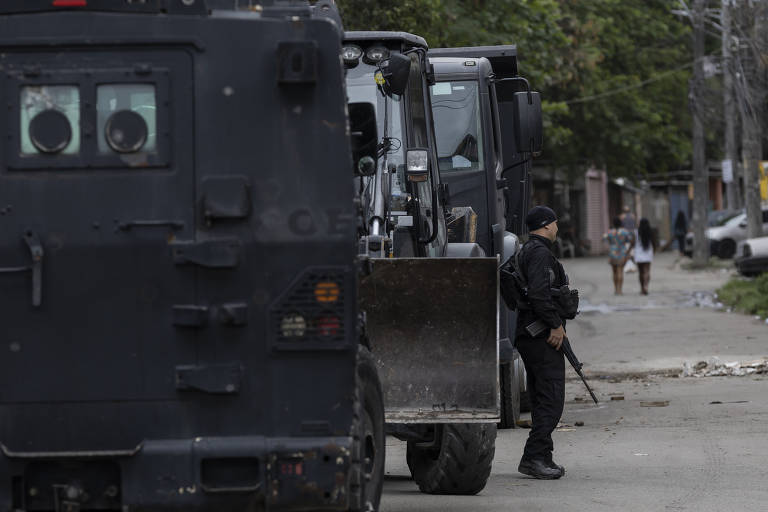 A imagem mostra uma operação policial em uma área urbana. Um policial armado está em pé ao lado de um veículo blindado preto, enquanto outro veículo pesado está estacionado próximo. Ao fundo, duas pessoas caminham pela rua. A cena ocorre em uma área residencial com árvores e casas ao redor