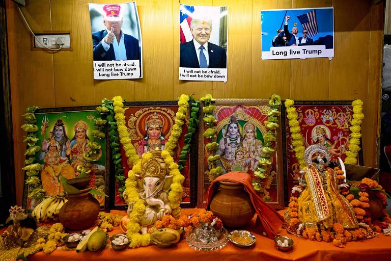 A imagem mostra um altar decorado com várias imagens religiosas hindus adornadas com guirlandas de flores amarelas. Na parte superior do altar, há três fotos de Donald Trump com textos em inglês. A primeira foto à esquerda tem o texto 'I will not be afraid, I will not be afraid, Long live Trump'. A foto central mostra Trump sorrindo, e a terceira foto à direita tem o texto 'Long live Trump'. O altar também contém diversos itens de oferenda, como frutas, potes de barro, e outros objetos decorativos.