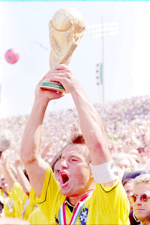 Imagem de um jogador de futebol vestindo uma camisa amarela e segurando um troféu acima da cabeça. Ele está comemorando, com a boca aberta em um grito de alegria. Há uma multidão ao fundo, também celebrando. O jogador usa uma faixa de capitão no braço esquerdo e uma medalha no pescoço.