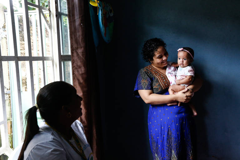 A imagem mostra uma mulher segurando um bebê em seus braços dentro de uma casa. A mulher está vestindo um vestido azul com detalhes dourados e o bebê está usando uma roupa clara com uma faixa rosa na cabeça. À esquerda, uma outra mulher está sentada perto de uma janela com grades, vestindo uma camiseta branca. A sala tem paredes escuras e uma cortina marrom pendurada na janela.