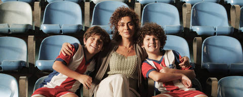 PEDAÇO DE MIM. (L to R) Theo Almeida as Mateus,  Juliana Paes as Liana, Enzo Diniz as Marcos in Pedaço de Mim. Cr. Marcos Serra Lima / Netflix © 2024 *** Local Caption *** ulher sentada em uma arquibancada, com um menino de cada lado. Os meninos estão usando uniformes de futebol com listras verticais vermelhas e brancas, meias azuis e chuteiras