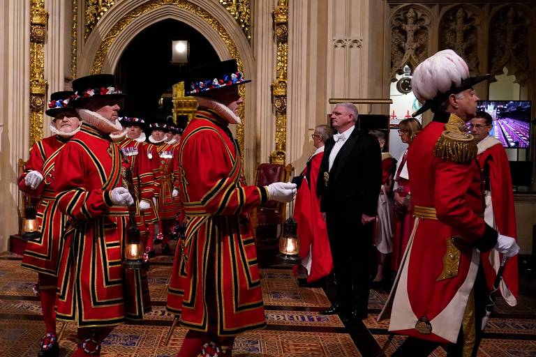 A imagem mostra uma cerimônia formal com guardas vestindo uniformes tradicionais vermelhos e pretos, com chapéus altos e decorados. Eles estão em um ambiente luxuoso com arquitetura gótica, incluindo arcos e detalhes dourados. Há também um homem de terno preto e gravata borboleta branca presente na cena. O ambiente parece ser um salão ou catedral com piso decorado.