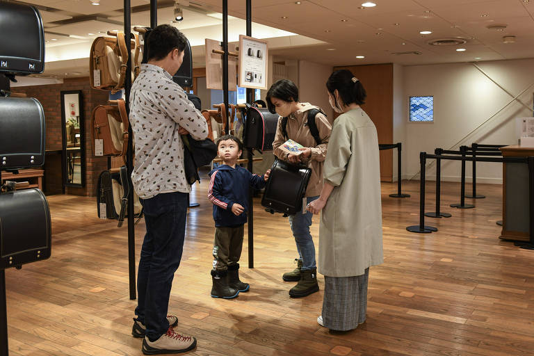 A imagem mostra uma família em uma loja de mochilas. Um homem está de pé com os braços cruzados, olhando para uma criança que está ao seu lado. A criança, vestida com uma jaqueta azul e botas, olha para uma mulher que segura uma mochila preta. Outra mulher, vestida com um casaco claro, também está presente. A loja tem piso de madeira e várias mochilas penduradas em expositores.