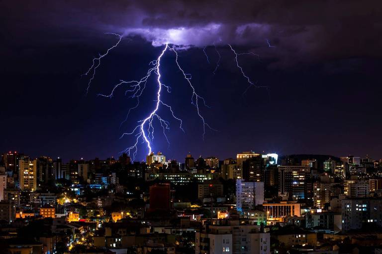 A imagem mostra um relâmpago iluminando o céu noturno sobre uma cidade. O relâmpago é brilhante e ramificado, descendo de nuvens escuras. A cidade abaixo está iluminada com luzes de edifícios e ruas, criando um contraste com o céu escuro