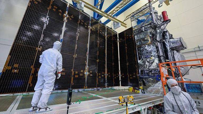 A imagem mostra dois técnicos usando trajes de proteção completos, incluindo máscaras e luvas, inspecionando um grande satélite em uma sala limpa. O satélite possui painéis solares grandes e está suspenso por uma estrutura metálica. Há equipamentos e ferramentas ao redor, e a sala tem um teto alto com vigas azuis