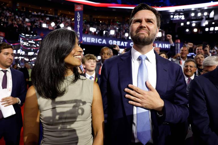 A imagem mostra um homem e uma mulher em um evento político. O homem está com a mão no peito e parece estar cantando ou recitando algo, enquanto a mulher ao seu lado sorri e olha para ele. Ao fundo, há uma multidão de pessoas e uma faixa com o texto 'AMERICA GREAT AGAIN!'.