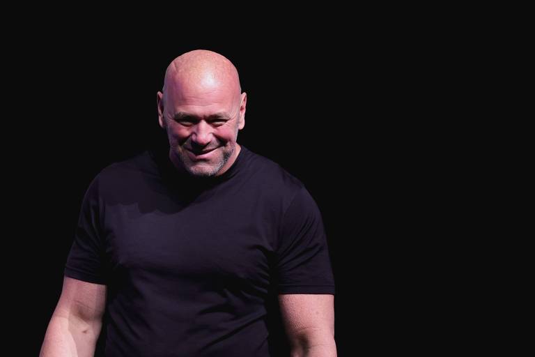 A imagem mostra Dana White, um homem careca sorrindo, vestindo uma camiseta preta. O fundo é completamente preto, destacando a figura do homem.