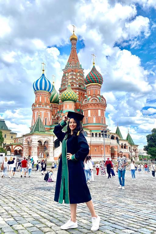 Uma mulher vestida com uma beca de formatura preta com detalhes verdes e um capelo está posando em frente à Catedral de São Basílio, em Moscou, Rússia. Ela está sorrindo e segurando o capelo com uma das mãos. Ao fundo, há várias pessoas caminhando na praça de paralelepípedos e o céu está parcialmente nublado com algumas áreas de céu azul.