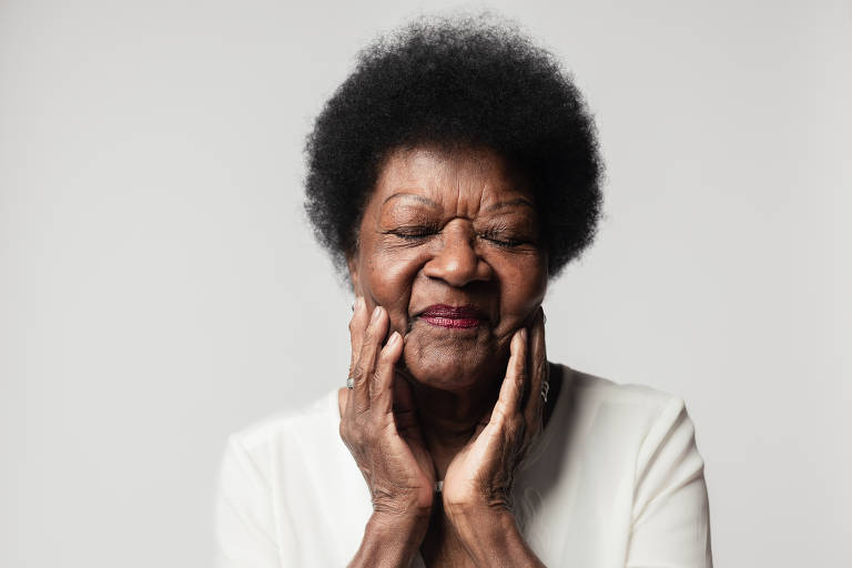 A imagem mostra uma mulher idosa, sorrindo suavemente enquanto toca seu rosto com as mãos. Ela está vestindo uma blusa clara e tem os olhos fechados, transmitindo uma expressão de alegria ou satisfação. O fundo é neutro e claro.