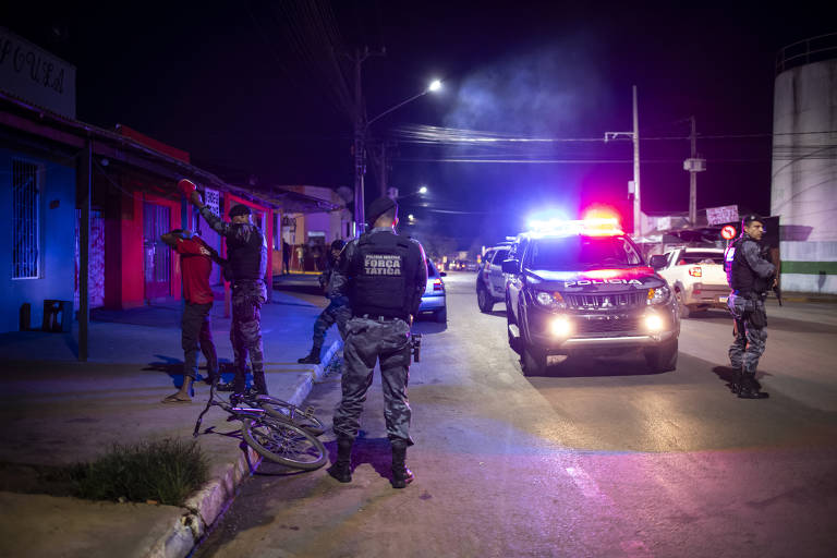 A imagem mostra uma operação policial noturna em uma rua. Policiais estão abordando dois homens que estão de pé contra a parede, com as mãos levantadas. Uma bicicleta está caída no chão ao lado deles. Um carro de polícia com luzes de emergência ligadas está estacionado na rua, iluminando a cena. Outros policiais estão presentes, observando a situação.