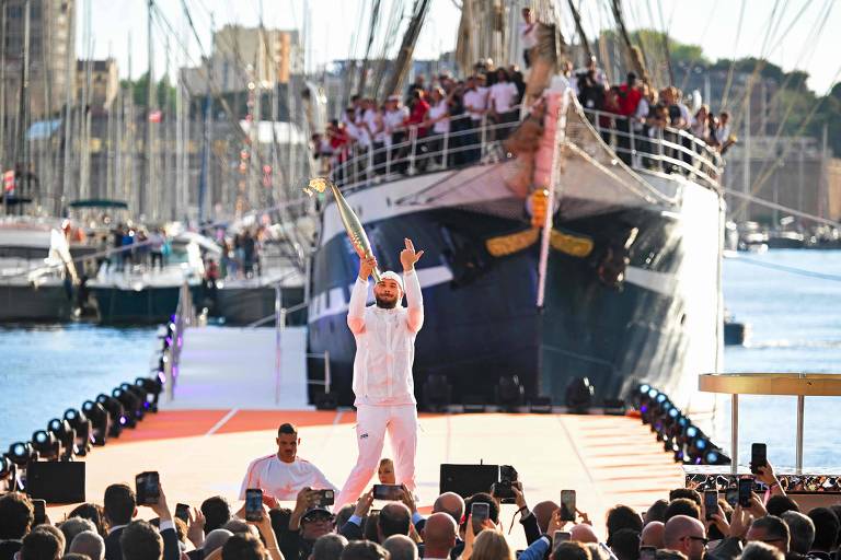 Imagem de um evento ao ar livre com uma grande multidão assistindo. No centro da imagem, uma pessoa vestida de branco está segurando um objeto acima da cabeça. Ao fundo, há um grande navio com várias pessoas a bordo, e o evento parece estar ocorrendo em um cais ou porto, com outros barcos visíveis ao fundo.