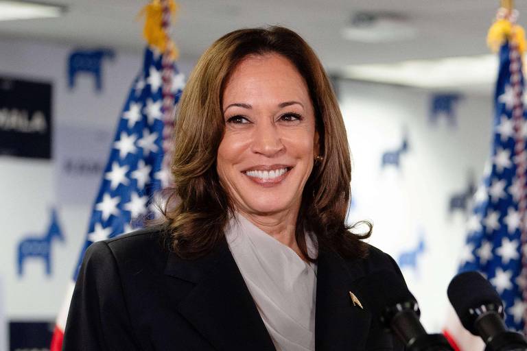 A imagem mostra uma mulher com cabelo castanho claro e liso, usando um terno escuro e uma blusa clara. Ela está sorrindo e parece estar em um evento, com bandeiras dos Estados Unidos ao fundo. Há microfones na frente dela, sugerindo que ela está prestes a falar ou já falou.