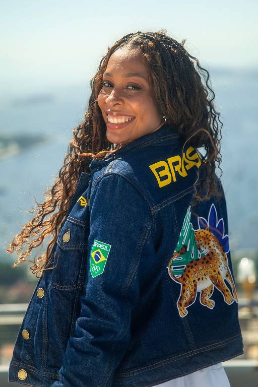 Mulher negra de cabelos compridos e cacheados está mostrando a parte traseira de jaqueta jeans e com a cabeça virada para a câmera. Lê-se "Brasil" em amarelo e tem o desenho de uma onça pintada bordada. Ela está em local ao ar livre, em dia de sol.