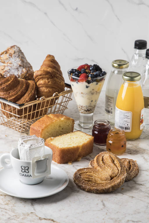 A imagem apresenta uma mesa com uma variedade de itens de café da manhã. Há uma cesta de metal com croissants e um pão doce. Ao lado, um copo com iogurte, granola e frutas vermelhas. Também estão presentes garrafas de água, suco de laranja e refrigerante. Uma xícara branca com o logotipo 'DAMA' contém sachês de chá. Pães e doces estão dispostos sobre uma superfície de mármore.