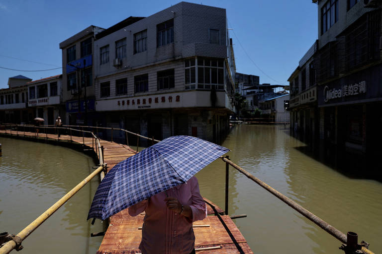 Pessoa segura guarda-sol enquanto caminha em uma ponte; as ruas do entorno estão alagadas, cobrindo parte das portas de edifícios