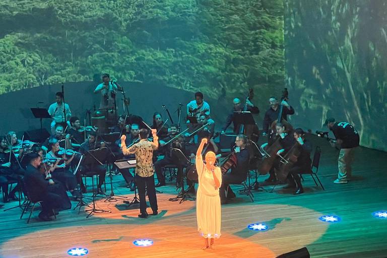 A imagem mostra uma apresentação musical ao vivo em um palco. No centro, uma cantora de vestido branco levanta o braço, enquanto um maestro, de costas, conduz uma orquestra composta por músicos com instrumentos variados. Ao fundo, há uma projeção de uma paisagem verde, possivelmente uma floresta. O palco é iluminado com luzes azuis e há um público visível em algumas partes da imagem.