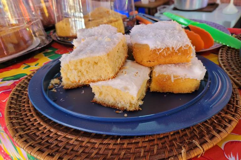 Um prato azul contém pedaços de bolo de coco cobertos com açúcar, dispostos de forma irregular. O fundo é uma mesa decorada com uma toalha colorida, e ao fundo, há outros itens de comida e utensílios.