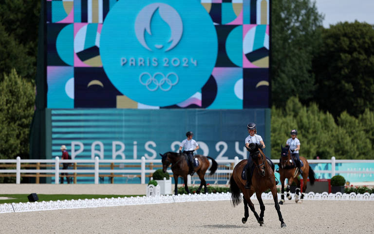 A imagem mostra um evento equestre com três cavaleiros montando seus cavalos em uma arena. Ao fundo, há um grande painel digital exibindo o logotipo dos Jogos Olímpicos de Paris 2024, que inclui a palavra 'PARIS' e os anéis olímpicos. O ambiente é ao ar livre, com árvores ao fundo e uma pista de areia.
