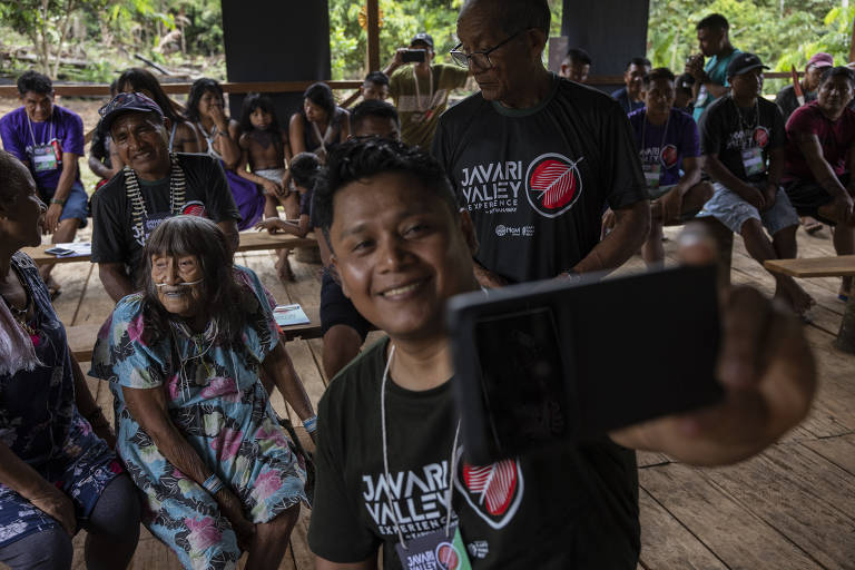 Varî Vãti Marubo está sentada cercada por outros indígenas, incluindo seu neto, que tira uma selfie com ela.