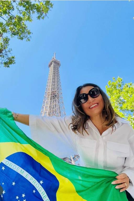 A imagem mostra uma mulher sorridente segurando a bandeira do Brasil em frente à Torre Eiffel. Ela está vestindo uma camisa branca e óculos escuros. O céu está claro e azul, com algumas folhas verdes visíveis ao redor.