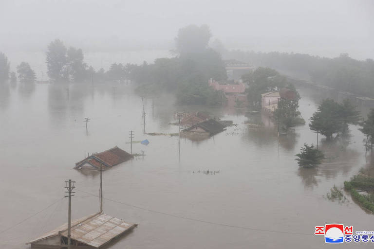 A imagem mostra uma área urbana severamente afetada por inundações, com várias casas submersas em água. A visibilidade é baixa devido à neblina, e árvores e postes de eletricidade são visíveis, parcialmente cobertos pela água. O cenário transmite uma sensação de desolação e gravidade da situação.