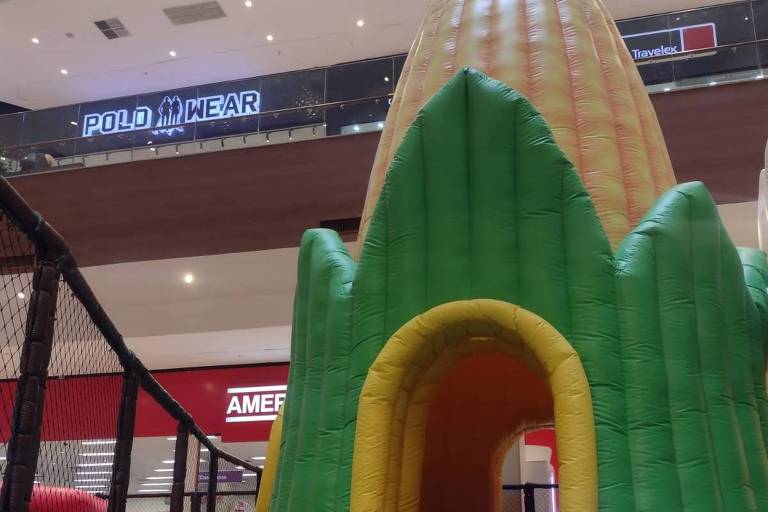 A imagem mostra um parque inflável em um shopping. O parque tem a forma de um grande milho, com cores verde e amarelo. Na frente, há uma entrada em forma de arco. O chão está coberto por uma variedade de bolinhas coloridas, predominantemente verdes, brancas e amarelas. Ao fundo, é possível ver uma loja com o nome 'POLD WEAR' e outra com o nome 'AMERIC'.