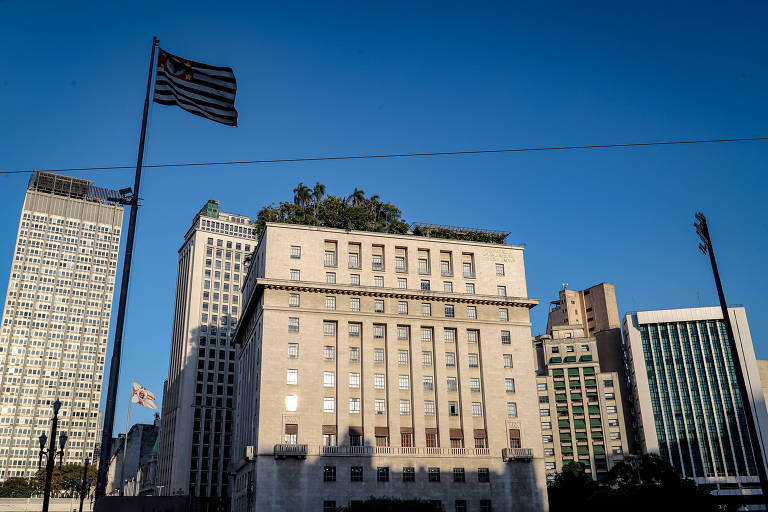 Imagem mostra Edifício Matarazzo, também conhecido como Palácio do Anhangabaú, a sede da prefeitura da cidade de São Paulo. O prédio é de estilo clássico e está em primeiro plano. Uma bandeira do estado de São Paulo está hasteada à esquerda