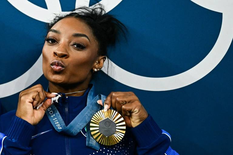 Uma atleta está posando com uma medalha de ouro ao redor do pescoço. Ela faz uma expressão de beijo, com os lábios levemente arredondados. A atleta usa um agasalho azul com detalhes brancos e tem cabelo preso. O fundo é azul com os anéis olímpicos em destaque.