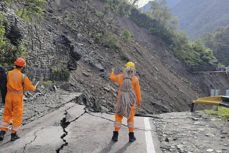 A imagem mostra um deslizamento de terra ao lado de uma estrada. Dois trabalhadores, vestidos com uniformes laranja e capacetes, estão observando a área afetada. O solo está instável e há uma fissura visível na estrada. Ao fundo, é possível ver vegetação e uma encosta montanhosa.