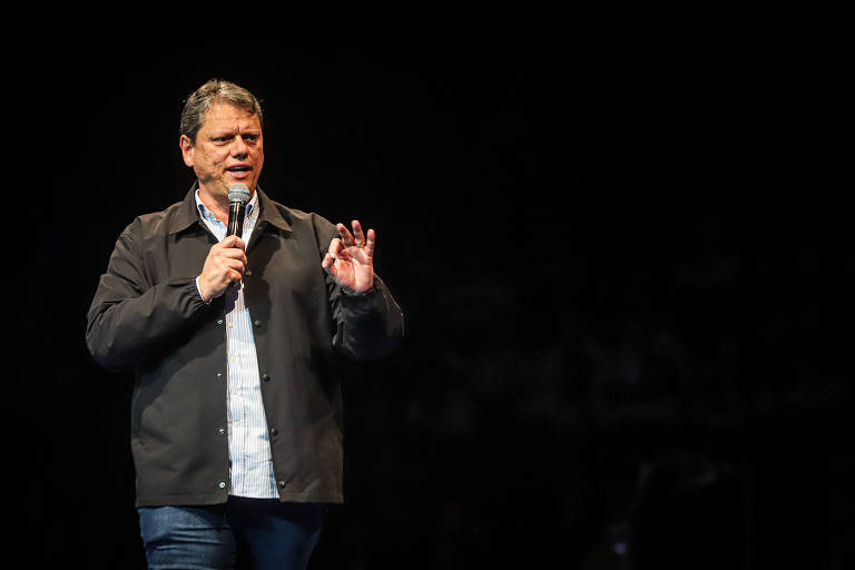 Um homem está em um palco, segurando um microfone e falando para a audiência. Ele usa uma jaqueta escura sobre uma camisa clara e jeans. O fundo é escuro, com a plateia visível em sombras.