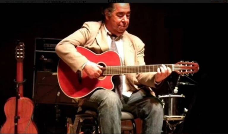 Homem sentado em um palco, tocando um violão acústico vermelho. Ele está vestido com um paletó claro e uma camiseta escura. Ao fundo, há um violão e um tambor. O ambiente parece ser um show ou apresentação musical