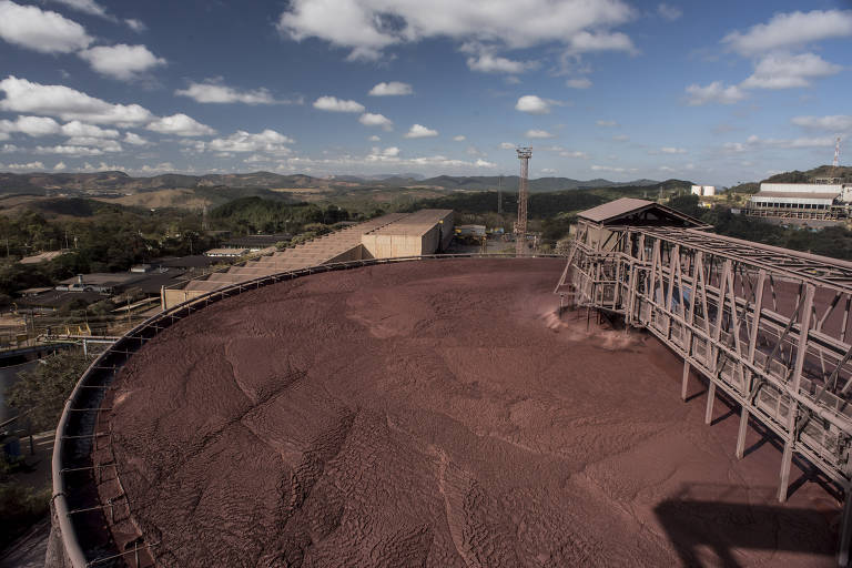 A imagem mostra uma grande área de uma mina de minério, com um depósito de rejeitos de cor avermelhada em primeiro plano. Ao fundo, há uma paisagem montanhosa com nuvens dispersas no céu. Estruturas industriais, como um galpão e torres, são visíveis, indicando a presença de atividades mineradoras na região.