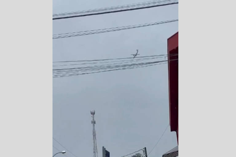 A imagem mostra um avião voando em um céu nublado, com fios de eletricidade visíveis na parte superior. No fundo, há uma estrutura de antena e um edifício vermelho à direita.