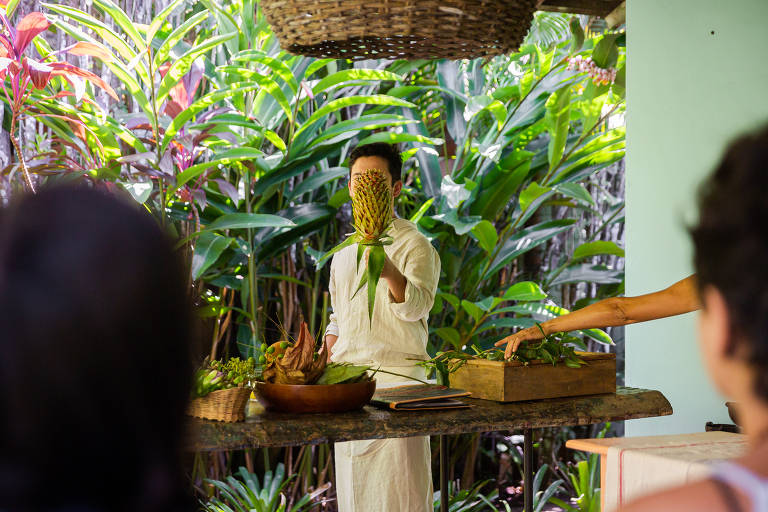 A imagem mostra uma pessoa em pé, segurando um abacaxi, em um ambiente tropical com muitas plantas verdes ao fundo. A pessoa está vestida com uma camisa branca e está em frente a uma mesa de madeira, onde há uma tigela com folhas e frutas. À direita, outra pessoa está estendendo a mão em direção à mesa. O cenário é iluminado e parece ser um espaço ao ar livre.