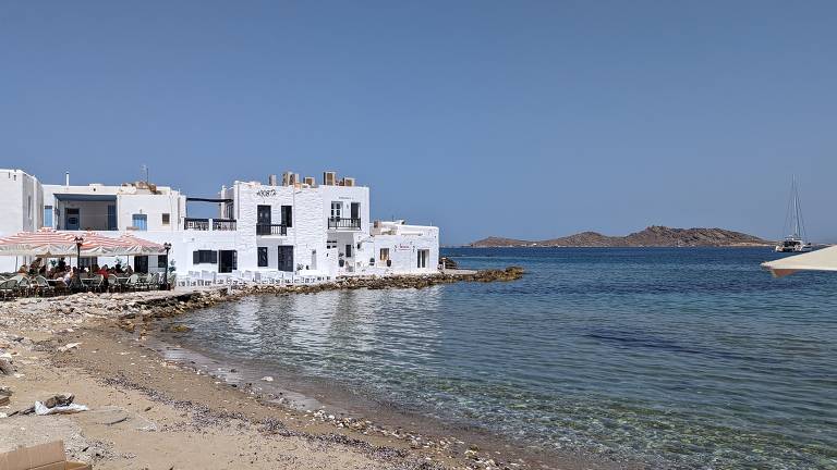 A imagem mostra uma praia com areia e pedras, onde há um restaurante à beira-mar com mesas e cadeiras. O restaurante possui toldos listrados em vermelho e branco. Ao fundo, há edifícios brancos com janelas azuis e uma vista para o mar, onde se pode ver uma pequena ilha e um barco à vela. O céu está limpo e azul.