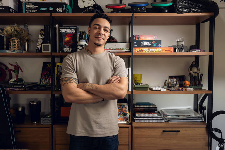 Um homem de pele clara, com cabelo curto e tatuagens nos braços, está em pé com os braços cruzados em frente a uma estante. Ele usa uma camiseta cinza e sorri levemente. A estante ao fundo contém diversos objetos, incluindo livros, jogos e itens decorativos.