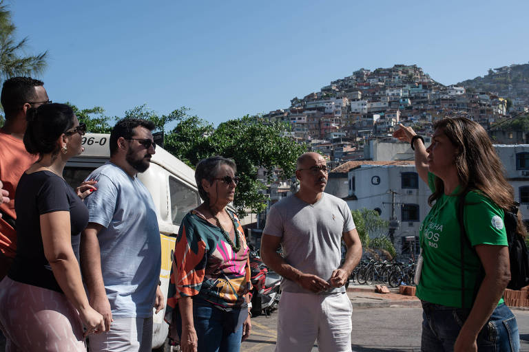 Um grupo de seis pessoas está em pé em uma área externa, olhando para uma colina com casas empilhadas. Uma mulher à direita, vestindo uma camiseta verde, está apontando para a colina enquanto fala. As outras cinco pessoas estão atentas, algumas com expressões de interesse. O céu está claro e ensolarado, e ao fundo, há uma vista de uma comunidade com muitas casas.