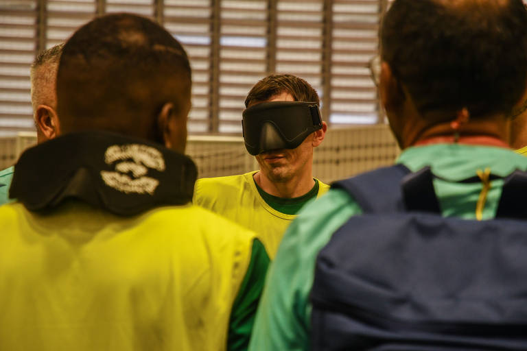 Foto de Karime Xavier mostra Ricardinho com a venda dos jogadores. Ele é um homem branco, com cabelos curtos e pretos. Ricardinho é visto entre os ombros de dois jogadores da seleção. Pela imagem é possível ver que ele está com o colete amarelo da seleção.