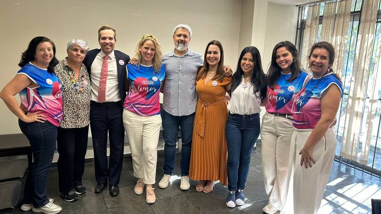 Um grupo de nove pessoas posando para a foto em um ambiente interno. Algumas pessoas estão usando camisetas coloridas com a frase 'Mutirão Amor'. O grupo inclui mulheres e homens, com diferentes estilos de cabelo e roupas. O fundo apresenta janelas com cortinas, permitindo a entrada de luz natural.