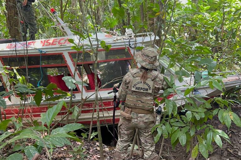 A imagem mostra um barco encalhado em uma área densa de floresta. O barco é de cor branca com detalhes em vermelho e possui a inscrição 'EMERG YSEC' na parte superior. Uma mulher vestindo um uniforme camuflado da Polícia Federal, com a inscrição 'NEPOM' nas costas, está de costas, observando o barco. Ao fundo, há vegetação densa, incluindo folhas verdes e troncos de árvores.