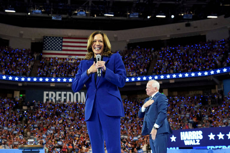 A imagem mostra uma mulher sorridente em um palanque, segurando um microfone. Ela está vestida com um terno azul e parece estar se dirigindo a uma grande multidão. Ao fundo, há uma bandeira dos Estados Unidos e uma plateia iluminada com luzes azuis. Um homem de terno escuro está ao lado dela, com a mão sobre o coração, em um gesto de respeito.