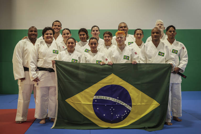 Um grupo de judocas posando em um tatame. Eles estão vestindo quimonos brancos e seguram uma bandeira do Brasil, que apresenta um fundo verde com um losango amarelo e um círculo azul. O grupo é composto por homens e mulheres, com diferentes características físicas. O ambiente é bem iluminado, com paredes verdes e um tatame azul e vermelho.