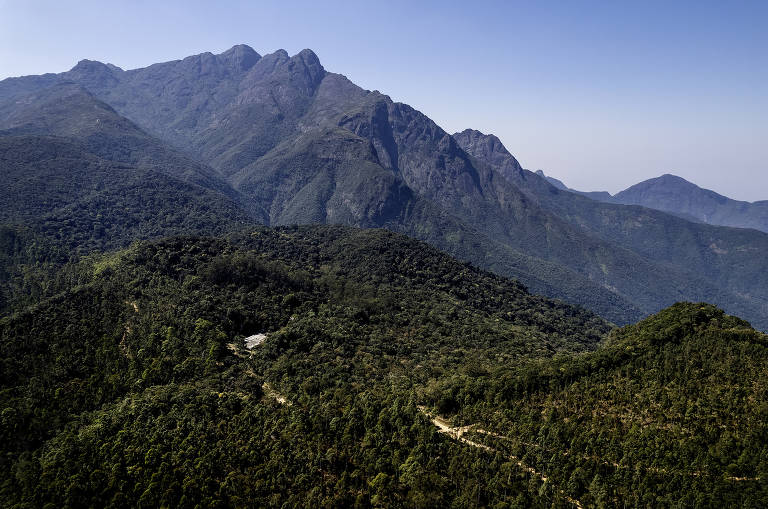 Imagem aérea de uma cadeia montanhosa com picos acentuados e vegetação densa. A montanha principal é alta e rochosa, cercada por colinas cobertas de árvores. Estradas sinuosas podem ser vistas ao longo da base das montanhas, levando a áreas mais baixas e abertas.