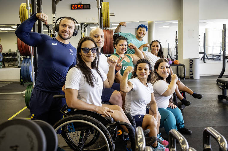 Um grupo de oito pessoas posando em uma academia. Algumas estão em pé e outras sentadas em cadeiras de rodas. Todos estão sorrindo e fazendo gestos de força com os braços. O ambiente é bem iluminado, com equipamentos de musculação ao fundo e pesos coloridos nas prateleiras. A temperatura no ambiente é de 25°C.