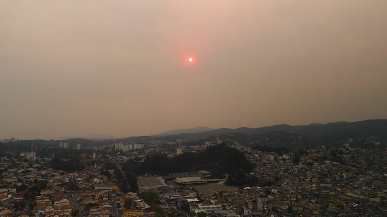 A imagem mostra um pôr do sol em uma cidade, com o céu coberto por uma névoa densa e poluída. O sol aparece como um ponto vermelho no horizonte, enquanto a cidade se estende abaixo, com várias construções e áreas urbanas visíveis. As montanhas estão ao fundo, parcialmente ocultas pela poluição.