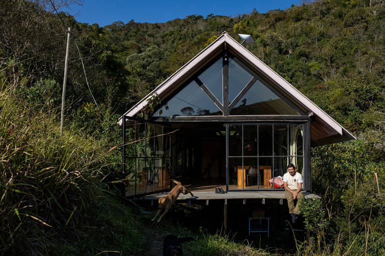 A imagem mostra uma casa moderna com um design de telhado em forma de pico, localizada em uma área verdejante. A estrutura é predominantemente de vidro e madeira, permitindo uma vista ampla do exterior. Na frente da casa, um homem está sentado em uma varanda, enquanto um cachorro está próximo a ele. O fundo é composto por colinas cobertas de vegetação.