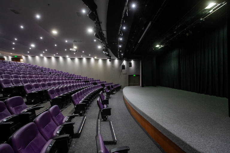 A imagem mostra o ambiente interno do Teatro Renaissance com assentos roxos dispostos em fileiras. O palco está à direita, com um piso coberto por carpete cinza e cortinas pretas ao fundo. O teto é iluminado por várias luzes, criando um ambiente bem iluminado. O teatro está vazio.