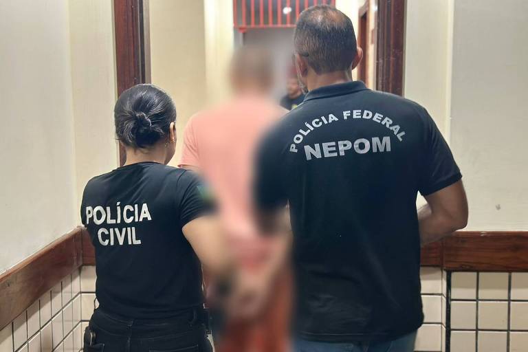 A imagem mostra dois agentes de segurança, um da Polícia Civil e outro da Polícia Federal, acompanhando uma pessoa detida em um corredor. O agente da Polícia Civil está usando uma camiseta preta com a inscrição 'POLÍCIA CIVIL' nas costas, enquanto o agente da Polícia Federal veste uma camiseta preta com a inscrição 'POLÍCIA FEDERAL NEPOM'. O ambiente é um corredor com paredes claras e azulejos na parte inferior.